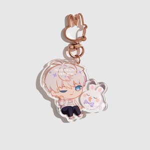 May include: An acrylic keychain featuring a cartoon character with white hair and blue eyes sitting next to a small white rabbit with a purple bowtie. The keychain has a rose gold rabbit-shaped clasp and includes the text Moonbuns sample.
