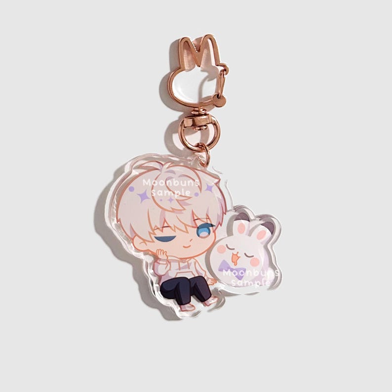 May include: An acrylic keychain featuring a cartoon character with white hair and blue eyes sitting next to a small white rabbit with a purple bowtie. The keychain has a rose gold rabbit-shaped clasp and includes the text Moonbuns sample.