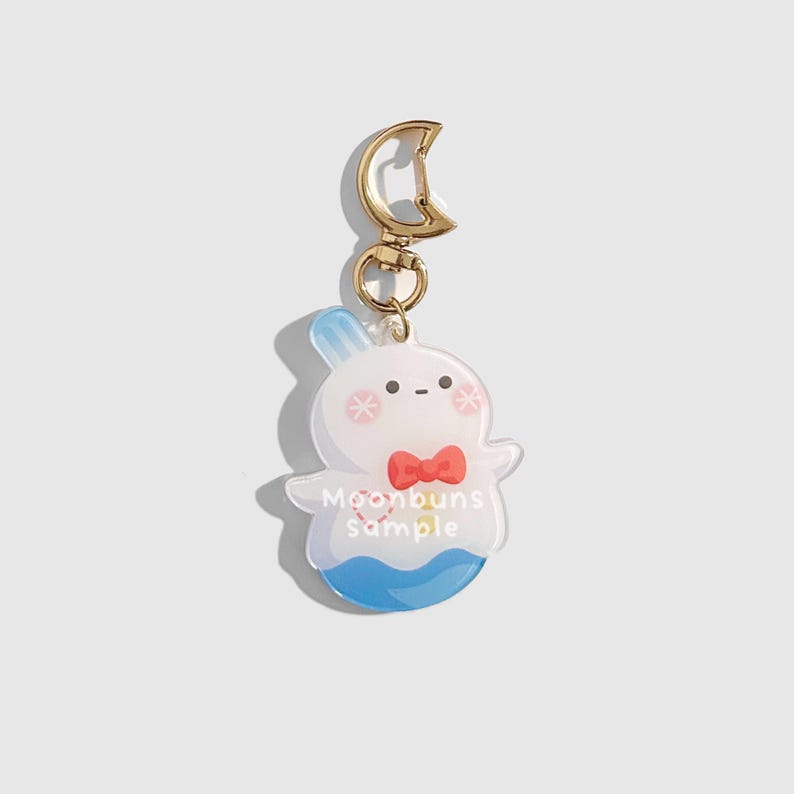 May include: A clear acrylic keychain featuring a cartoon bunny-like character with a red bow tie and blue accents. The keychain has a gold-colored clasp and a crescent-shaped charm. The text "Moonbuns sample" is printed on the keychain.