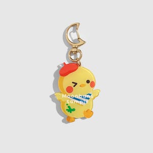 May include: A yellow chick-shaped keychain with a red beret, blue scarf, and green paint splat. The keychain has a gold-colored clasp and a small ring. The text "Moonbuns sample" is printed on the chick's body.
