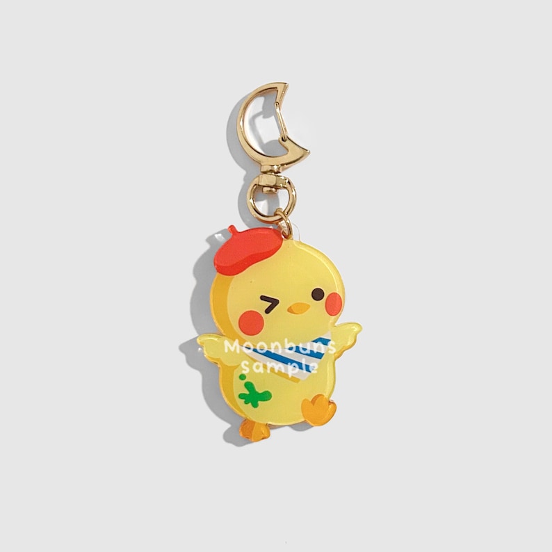 May include: A yellow chick-shaped keychain with a red beret, blue scarf, and green paint splat. The keychain has a gold-colored clasp and a small ring. The text "Moonbuns sample" is printed on the chick's body.