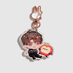 May include: An acrylic keychain featuring a cartoon character with brown hair and a dark outfit sitting next to a grumpy red apple character. The keychain has a rose gold rabbit-shaped clasp and includes the text Moonbuns sample on both figures.
