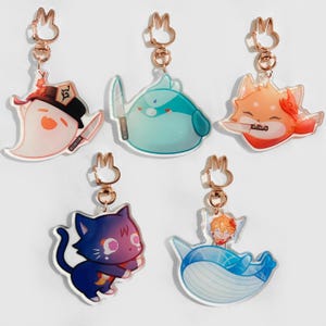 Puede incluir: Cinco llaveros de acrílico coloridos con anillos de metal dorado. Los llaveros presentan personajes de dibujos animados con diferentes expresiones y poses. Los personajes incluyen un fantasma, una mancha azul, un zorro, un gato negro y una ballena con una persona encima.