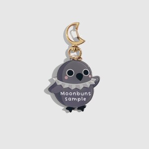 May include: A gray bird-shaped keychain with a gold-colored clasp. The bird has a white ruffled collar, pink cheeks, and the text "Moonbuns sample" on its belly. The keychain is a cute accessory.