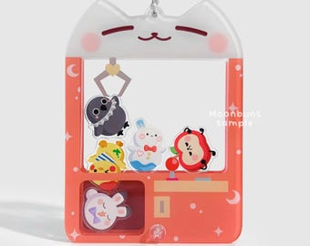 May include: A clear, cat-shaped keychain with a red claw machine design. The machine contains various cartoon characters, including a bird, a bunny, and a red apple. The text "Moonbuns sample" is visible on the side.
