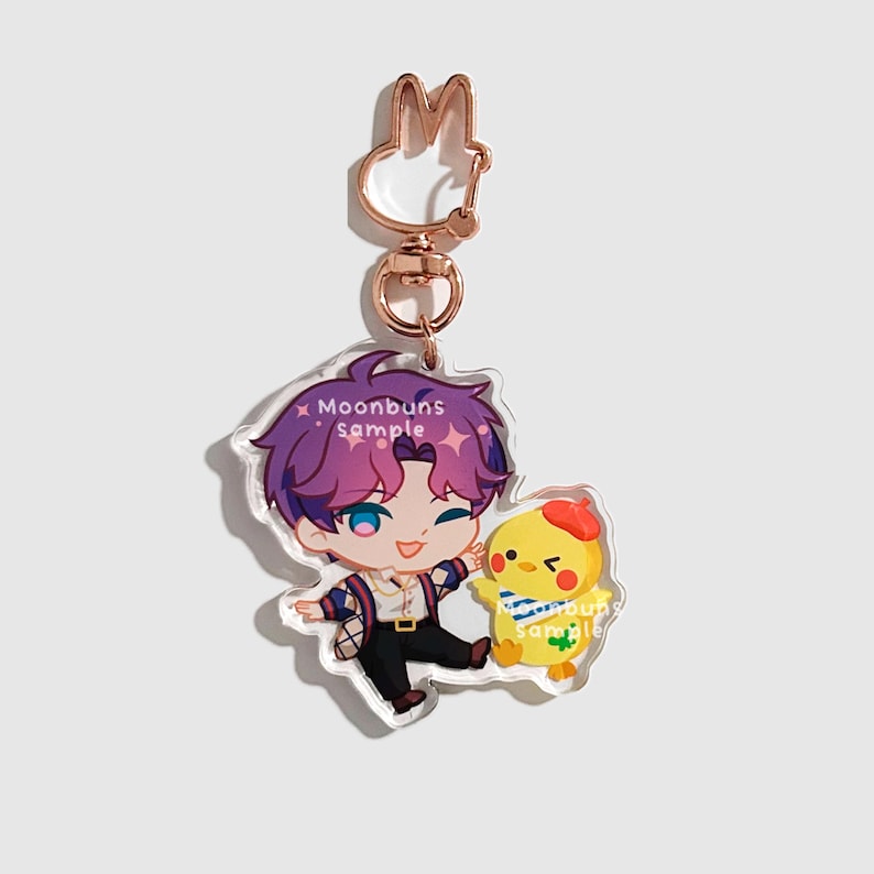 May include: An acrylic keychain featuring a cartoon character with purple hair and a yellow chick wearing a red beret. The character wears a white shirt and dark pants. The text Moonbuns sample is printed on the character and the chick. It has a rose gold rabbit-shaped clasp.