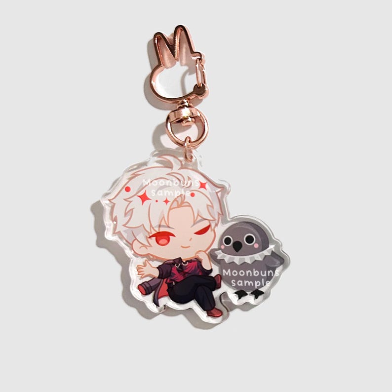 May include: An acrylic keychain featuring a cartoon character with white hair and red eyes, wearing a dark outfit, sitting next to a small gray bird-like creature. The charm is attached to a rose gold-colored metal clasp shaped like a rabbit head.