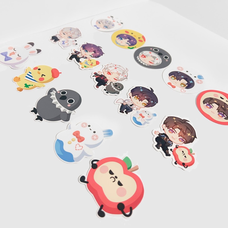 May include: A collection of colorful stickers featuring various cartoon characters and designs. The stickers include images of anime-style figures, animals, and food items like an apple. The stickers are arranged on a white surface.
