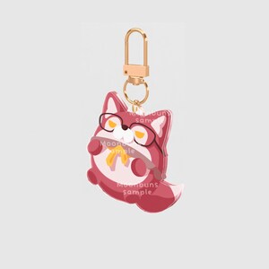 May include: A keychain featuring a stylized red and white fox character wearing black glasses and a yellow necktie. The fox has a rounded body and is attached to a gold-colored metal lobster clasp. The surface displays the text Moonbuns sample.