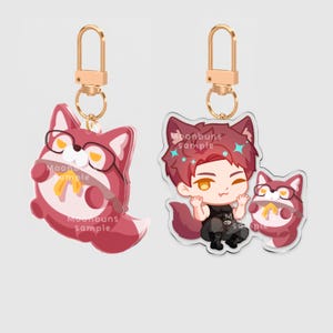 May include: Two acrylic keychains with gold-tone lobster clasps. One features a round, red, fox-like creature wearing glasses and a yellow bow. The other shows a red-haired character with fox ears next to the same fox creature, both with text overlays.