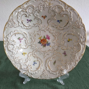Könnte beinhalten: Ein weißer Porzellanteller mit einem aufwendigen Relief-Rand und Goldverzierung. Die Mitte und die umliegenden Bereiche sind mit kleinen, handgemalten Blumensträußen in den Farben Orange, Rosa, Gelb, Blau und Violett verziert.