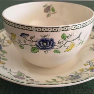 Puede incluir: Un juego de taza y platillo de cerámica color crema con un delicado patrón floral de flores azules, amarillas y moradas sobre tallos verdes. El borde de ambas piezas está decorado con una cenefa geométrica verde oscuro y una asa.