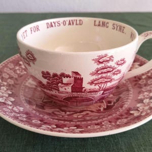 Könnte beinhalten: Ein cremefarbenes Tassen- und Untertassen-Set aus Keramik mit einem dunkelrosa Transferdruck-Muster aus Schloss, Brücke und Bäumen. Der innere Rand der Tasse ist mit dem Text YET FOR DAYS O'AVLD LANG SYNE in dunkelrosa Schrift versehen.