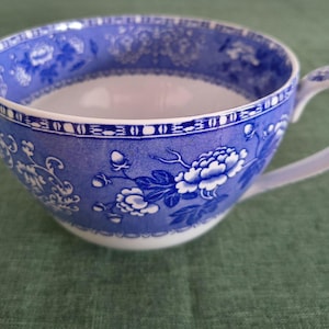 May include: A ceramic teacup featuring a white base with a vibrant blue floral pattern and intricate border designs. The cup has a curved handle and a wide, open bowl shape, showcasing detailed botanical illustrations in a deep blue hue.
