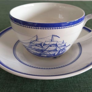 Puede incluir: Un juego de taza de té y platillo de porcelana blanca con una ilustración náutica azul de un velero. El borde de la taza y el borde del platillo están decorados con una banda azul y un delicado patrón festoneado en un color azul a juego.