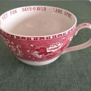 Puede incluir: Una taza grande, blanca, con un estampado floral rojo y las palabras "YET FOR DAYS O'AVLD LANG SYNE" impresas en el borde. La taza tiene un asa curva y está sobre un fondo verde.