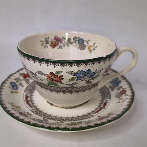 May include: A vintage teacup and saucer set. The cream-coloured cup and saucer feature a floral design in shades of red, blue, and orange. The cup has a green rim and handle, and the saucer has a matching border. A classic tea set.