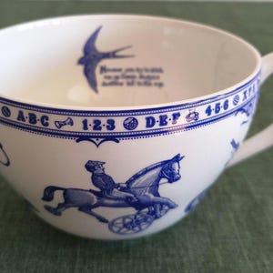 Puede incluir: Una taza de té de cerámica blanca con ilustraciones y texto en azul. El borde muestra una cenefa con letras, números e iconos. El cuerpo presenta un jinete en un carruaje de caballos. Dentro, un gráfico de un pájaro sobre texto impreso.