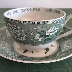 Könnte beinhalten: Ein grün-weißes Keramik-Tassenset mit Untertasse, verziert mit einem Blumenmuster und dem Text YET FOR DAYS O'AVLD LANG SYNE am inneren Rand. Die Tasse hat einen geschwungenen Henkel und die Untertasse zeigt ein passendes, detailliertes Pflanzenmotiv.