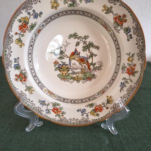 Puede incluir: Un plato redondo de cerámica con base color crema que presenta una ilustración central de dos pájaros coloridos posados en una rama de árbol cerca de una urna llena de flores. El borde está decorado con un patrón repetitivo de flores naranjas, amarillas y azules.
