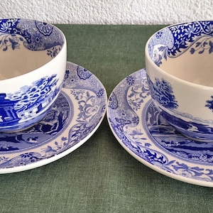 Puede incluir: Dos tazas y platillos a juego azules y blancos. Las tazas y los platillos tienen un diseño de paisaje detallado. Las tazas tienen un asa curva y se asientan sobre platillos a juego. El diseño incluye elementos florales y arquitectónicos.