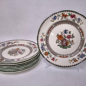 Könnte beinhalten: Ein Satz Vintage-Porzellanteller mit floralem Design. Die Teller haben einen weißen Hintergrund mit einem braunen, gemusterten Rand und einem grünen Rand. Die Mitte jedes Tellers zeigt ein farbenfrohes Blumenarrangement.