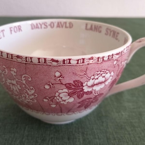 Puede incluir: Una taza de té de cerámica blanca con un patrón floral de color rosa oscuro en el exterior y un asa a juego. El borde interior tiene la inscripción 'IS YET FOR DAYS-O-AVLD LANG SYNE' en letras de color rosa oscuro.