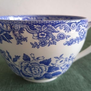 May include: A white ceramic teacup featuring a blue floral pattern. The design includes a detailed border of small flowers and leaves around the rim, with larger, prominent floral clusters decorating the body of the cup and a curved handle.