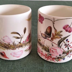 Puede incluir: Dos tazas de cerámica blanca con una detallada ilustración botánica que rodea el exterior. El diseño incluye flores rosas, bellotas marrones, hojas verdes y pequeñas bayas rojas, con un motivo floral en la parte superior de cada asa.