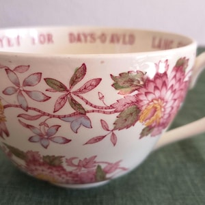 May include: A cream-coloured ceramic teacup featuring a pink and green floral pattern around the exterior. The interior rim displays printed text that reads "DAYS O AULD LANG SYNE" in a reddish-pink font. The cup has a curved handle on the right side.