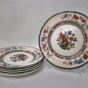 Puede incluir: Un juego de seis platos de cerámica blanca con un diseño floral. Cada plato presenta un ramo floral central, un borde estampado y un borde verde festoneado. Los platos están apilados y expuestos sobre un soporte transparente.