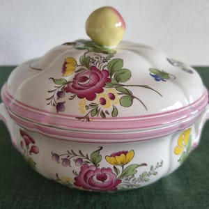Puede incluir: Una sopera de cerámica blanca con tapa, decorada con un patrón floral pintado a mano en tonos rosa, amarillo y verde. La tapa tiene un pequeño pomo con forma de fruta, y el cuenco cuenta con dos asas laterales y un borde de color rosa.