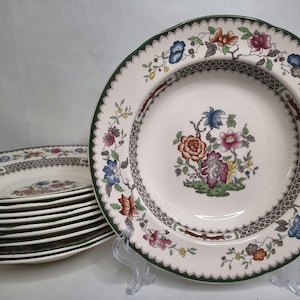 May include: A collection of cream-coloured ceramic plates with a floral pattern. The plates have a green scalloped edge, a central floral design, and a decorative border. The plates are stacked and displayed on a clear stand.