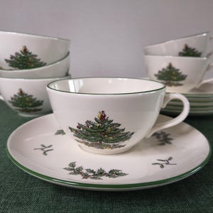 Puede incluir: Un juego de tazas de té y platillos de cerámica blanca con un gráfico de árbol de Navidad verde con adornos coloridos y un borde verde. Los platillos están decorados con pequeños patrones de hojas de acebo. Las tazas tienen un asa lateral.