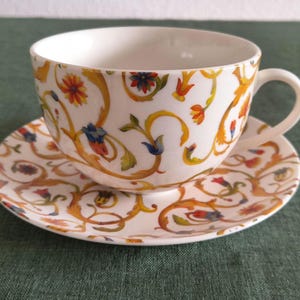 Könnte beinhalten: Eine weiße Porzellantasse mit passender Untertasse, verziert mit einem Muster aus goldgelben Ranken, grünen Blättern sowie kleinen roten und blauen Blüten. Die Tasse besitzt einen geschwungenen Henkel und einen glatten, abgerundeten Rand.
