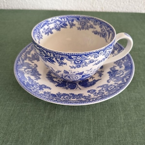 Könnte beinhalten: Eine weiße Teetasse mit passender Untertasse, verziert mit einem detaillierten blauen Blumenmuster. Das Design umfasst komplexe Blüten, Blätter und dekorative Schnörkelränder, die sich um die Außenseite der Tasse und den Rand der Untertasse ziehen.