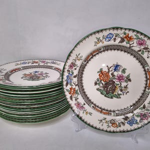 Op de afbeelding: Een set vintage keramische borden met een bloemenontwerp. De borden hebben een witte achtergrond met een bruine rand en een groene rand. Het bloemenontwerp bevat oranje, roze, blauwe en bruine bloemen. De borden zijn opgestapeld.