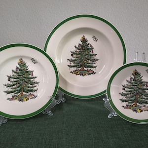 May include: Three cream-coloured ceramic plates of varying sizes, each featuring a green rim and a central illustration of a decorated Christmas tree with colourful ornaments and wrapped gifts at the base, displayed on clear plastic stands.
