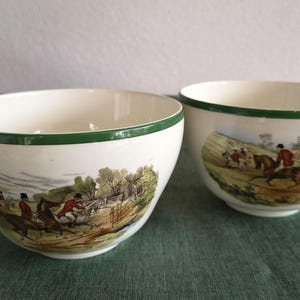 Könnte beinhalten: Zwei cremefarbene Keramik-Teetassen mit grünem Rand und braunem Henkel. Jede Tasse zeigt eine detaillierte Außenszene mit Reitern in einer Landschaft. Die Tassen haben eine breite, abgerundete Form und eine glatte, glänzende Oberfläche.