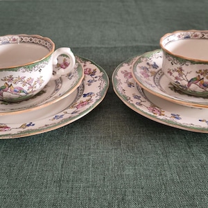 Puede incluir: Dos juegos de tazas de té y platillos de porcelana, cada uno acompañado de un plato a juego. La cerámica blanca presenta un delicado patrón de pájaros coloridos sobre ramas florecidas, con bordes verdes y detalles en oro.