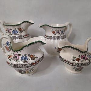 Könnte beinhalten: Vier weiße Keramik-Sahnekännchen mit floralen Mustern in Blau, Rosa, Orange und Braun. Jedes Kännchen hat einen gewellten Rand mit grünem Rand und einen Henkel. Die Kännchen sind unterschiedlich groß.