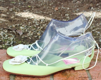 May include: A pair of light green leather shoes with white flower embellishments. The shoes have a clear plastic overlay and white laces.