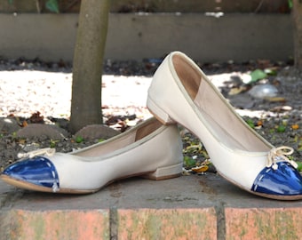 May include: Two pairs of off-white ballet flats with blue patent leather toes. The shoes have a bow on the top of the foot.