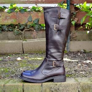 May include: Brown leather knee-high boots with buckle accents. The boots have a low block heel and a pointed toe.