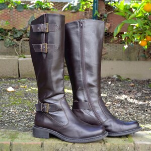 May include: A pair of brown leather knee-high boots with buckles and zippers. One boot has a buckle detail on the side, while the other has a zipper detail.