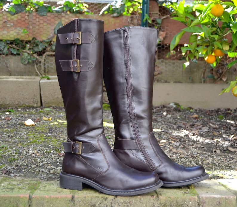 May include: A pair of brown leather knee-high boots with buckles and zippers. One boot has a buckle detail on the side, while the other has a zipper detail.