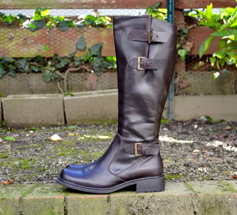 May include: Brown leather knee-high boots with buckle accents. The boots have a low block heel and a pointed toe.