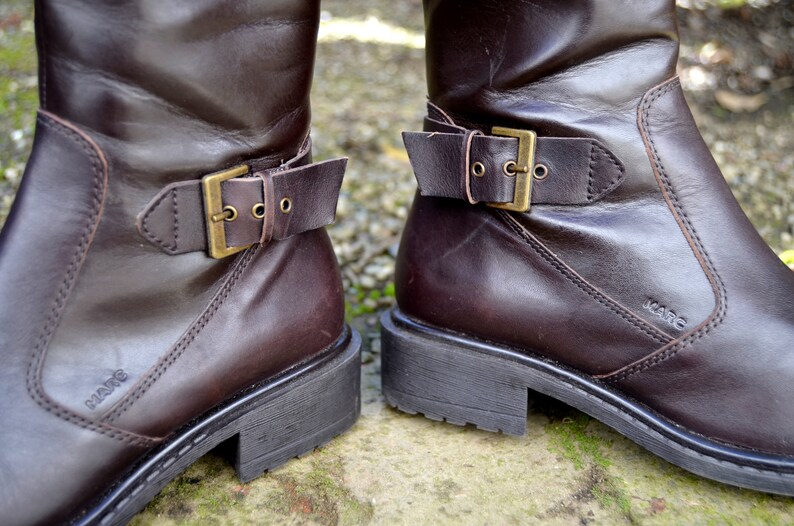 May include: A pair of brown leather boots with buckles. The boots have a stitched seam on the side and the brand name "Kare" is visible on the right boot.