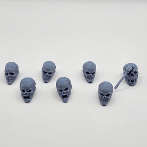 Puede incluir: Una colección de pequeñas figuras de calaveras grises con detalles. Una calavera está emparejada con un martillo en miniatura. Las calaveras están dispuestas sobre una superficie blanca, lo que sugiere un tema de terror o fantasía.