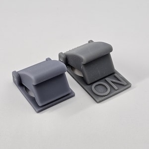 Op de afbeelding: Twee grijze plastic schakelaars, een met het woord "ON" gedrukt op de basis. De schakelaars zijn ontworpen om op een oppervlak te worden gemonteerd.